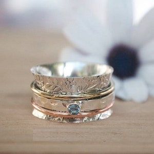 Könnte beinhalten: Ein silberner Drehring mit einem blauen Edelstein und einem Blumenmuster. Der Ring besteht aus drei Bändern, einem silbernen, einem goldenen und einem kupfernen.