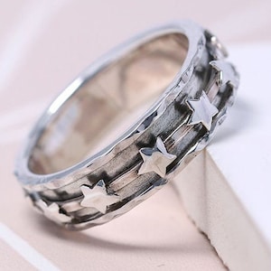May include: A silver ring with a textured band and five star-shaped accents. The stars are arranged in a staggered pattern around the band.