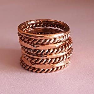 K&ouml;nnte beinhalten: Ein Stapel aus f&uuml;nf Kupferringen mit einem geflochtenen Design. Die Ringe sind &uuml;bereinander gestapelt und haben eine glatte, polierte Oberfl&auml;che.