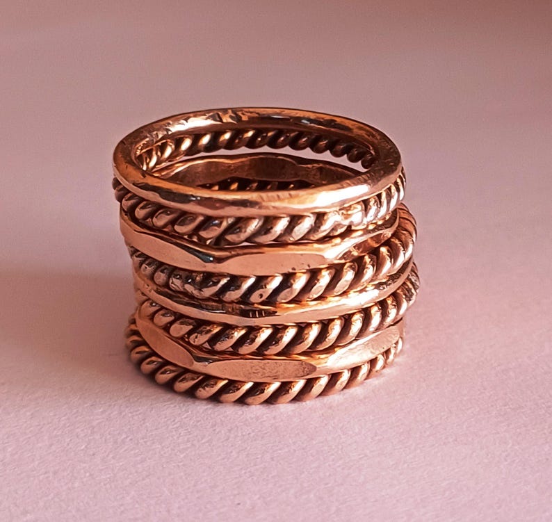 K&ouml;nnte beinhalten: Ein Stapel aus f&uuml;nf Kupferringen mit einem geflochtenen Design. Die Ringe sind &uuml;bereinander gestapelt und haben eine glatte, polierte Oberfl&auml;che.