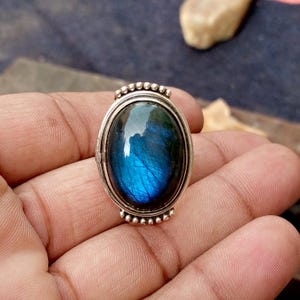 May include: Silver ring with an oval, iridescent blue gemstone. The gemstone displays a vibrant play of colour, with blue and green hues. The ring has a detailed silver setting with a beaded design around the gemstone.