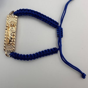 Può includere: Un braccialetto intrecciato blu con un ciondolo dorato raffigurante una figura religiosa. Il braccialetto ha un nodo scorrevole per regolare la dimensione.