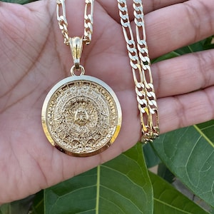 May include: A gold chain necklace with a large, round pendant featuring an intricate design. The pendant has a textured surface and appears to be made of metal.