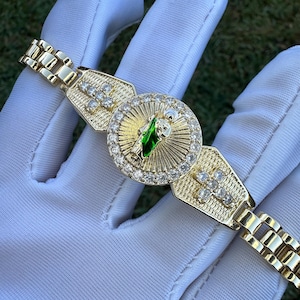 May include: A gold bracelet with a round charm featuring a religious figure with a green gem. The charm is surrounded by clear stones. The bracelet has a chain link design.