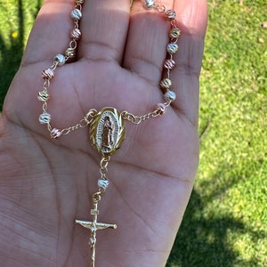 May include: A gold-tone rosary bracelet with a pendant featuring the Virgin Mary and a crucifix. The beads are a mix of gold, silver, and rose gold.