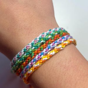 May include: A collection of colourful friendship bracelets. Each bracelet is made of woven threads in various colours, including green, orange, purple, and yellow. The bracelets are worn on a wrist.