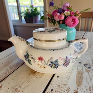 May include: White ceramic teapot with a floral design in pink, blue, and green. The teapot has a lid with a gold rim and a matching floral pattern. The teapot is on a distressed white wooden table.