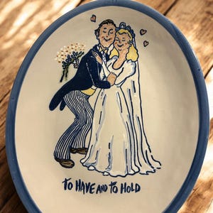 May include: A ceramic plate with a blue rim featuring a hand-painted wedding scene. The illustration depicts a couple embracing, with the text "TO HAVE AND TO HOLD" below. The plate has a cream-colored background.