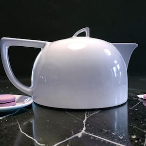 May include: A white ceramic teapot with a curved handle and a small spout. A small white plate with a purple macaron is to the left of the teapot. The teapot is on a black marble surface.