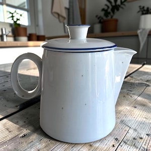 May include: A white ceramic teapot with a speckled finish and a blue rim on the lid. The teapot features a circular handle on the left side, a short spout on the right, and a matching lid with a small knob handle on top. The surface is smooth.