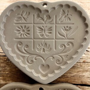 Puede incluir: Molde gris en forma de coraz&oacute;n con dise&ntilde;os florales y de mariposas en relieve. El molde tiene un borde festoneado y un peque&ntilde;o agujero para colgar. El dise&ntilde;o incluye una cuadr&iacute;cula de nueve cuadrados con varios patrones florales en relieve.