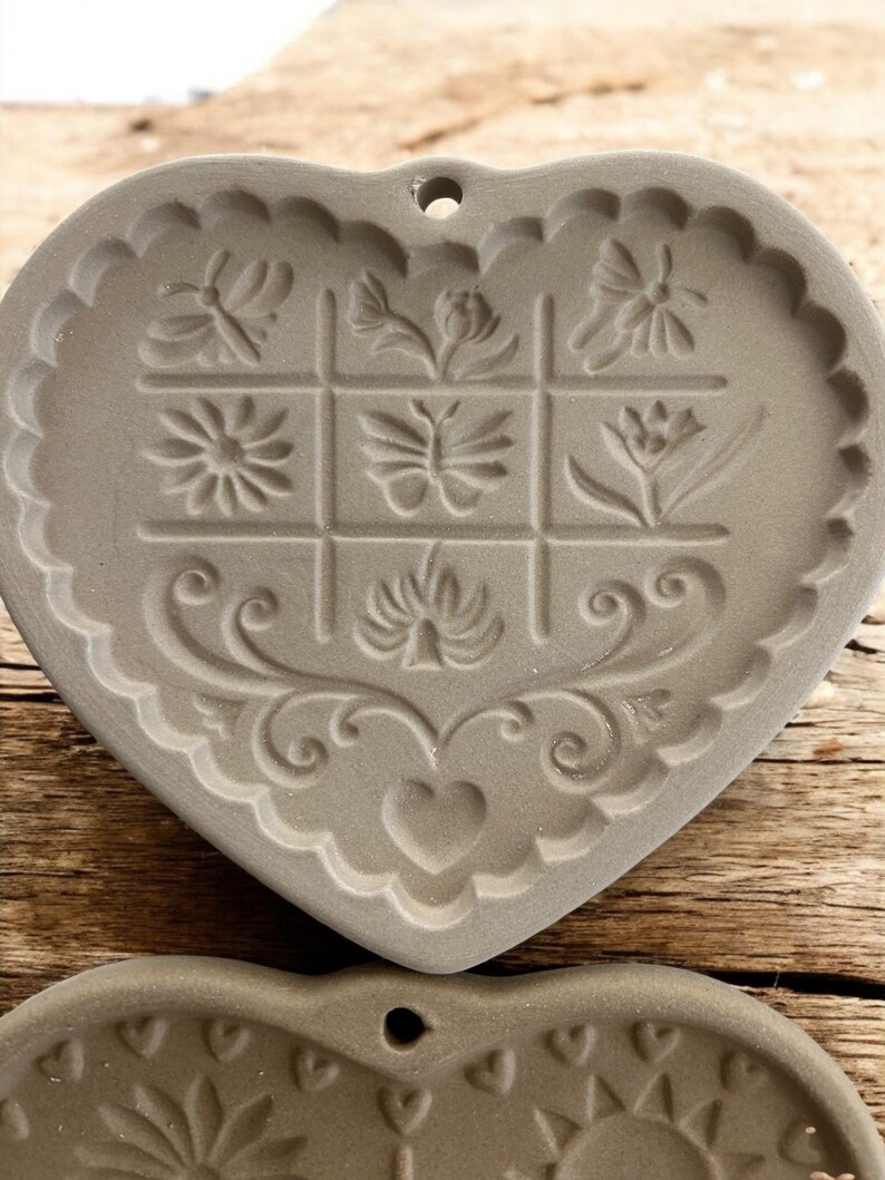 Puede incluir: Molde gris en forma de coraz&oacute;n con dise&ntilde;os florales y de mariposas en relieve. El molde tiene un borde festoneado y un peque&ntilde;o agujero para colgar. El dise&ntilde;o incluye una cuadr&iacute;cula de nueve cuadrados con varios patrones florales en relieve.