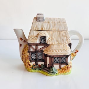 May include: A ceramic teapot shaped like a cottage with a thatched roof, a chimney, and timber-framed walls. The exterior features painted details of lattice windows, green shrubbery, and autumn-colored foliage around the base of the structure.