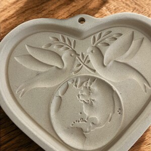 Puede incluir: Un molde de cer&aacute;mica gris claro en forma de coraz&oacute;n con dos palomas sosteniendo ramas de olivo sobre un globo terr&aacute;queo. El molde tiene un peque&ntilde;o agujero en la parte superior para colgarlo. El dise&ntilde;o est&aacute; en relieve, creando una superficie texturizada.