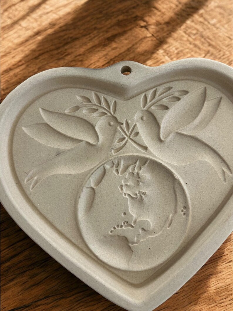 Puede incluir: Un molde de cer&aacute;mica gris claro en forma de coraz&oacute;n con dos palomas sosteniendo ramas de olivo sobre un globo terr&aacute;queo. El molde tiene un peque&ntilde;o agujero en la parte superior para colgarlo. El dise&ntilde;o est&aacute; en relieve, creando una superficie texturizada.