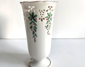 May include: A white porcelain vase featuring a scalloped rim with intricate cut-out lattice detailing. The body is decorated with green holly leaves and red berries. The base and the top rim are finished with a thin gold metallic trim.