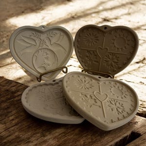 Puede incluir: Cuatro posavasos de cer&aacute;mica en forma de coraz&oacute;n con dise&ntilde;os en relieve. Uno presenta palomas y un globo terr&aacute;queo, mientras que otros muestran soles, copos de nieve y motivos florales. Cada posavasos tiene un peque&ntilde;o agujero en la parte superior. Los posavasos son beige.