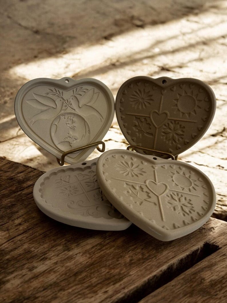 Puede incluir: Cuatro posavasos de cer&aacute;mica en forma de coraz&oacute;n con dise&ntilde;os en relieve. Uno presenta palomas y un globo terr&aacute;queo, mientras que otros muestran soles, copos de nieve y motivos florales. Cada posavasos tiene un peque&ntilde;o agujero en la parte superior. Los posavasos son beige.