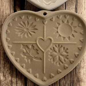 Puede incluir: Salvamanteles de cer&aacute;mica en forma de coraz&oacute;n, de color beige, con dise&ntilde;os de temporada en relieve. Presenta un sol, un copo de nieve, una hoja y una flor, con un coraz&oacute;n central y peque&ntilde;os corazones alrededor del borde.