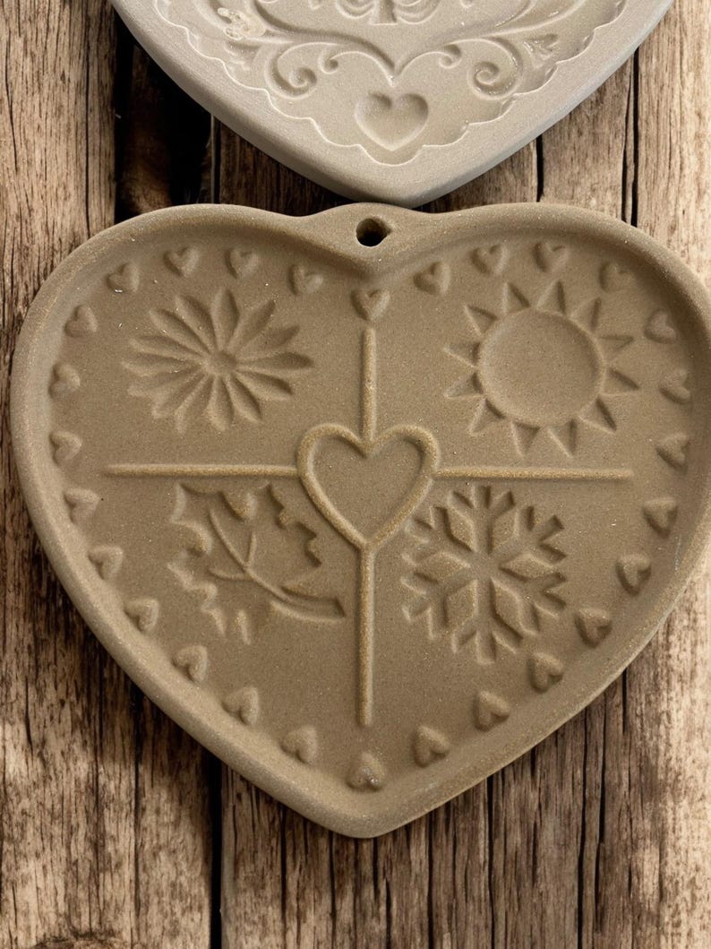 Puede incluir: Salvamanteles de cer&aacute;mica en forma de coraz&oacute;n, de color beige, con dise&ntilde;os de temporada en relieve. Presenta un sol, un copo de nieve, una hoja y una flor, con un coraz&oacute;n central y peque&ntilde;os corazones alrededor del borde.