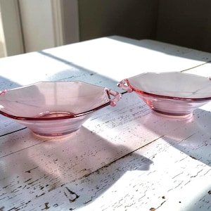 May include: Two matching pink glass bowls with a shallow, octagonal shape and small handles on opposite sides. The translucent glass features a smooth, polished finish and a wide rim, creating a delicate appearance for these serving dishes.