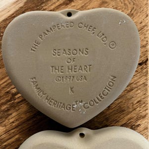 Puede incluir: Dos adornos de cer&aacute;mica en forma de coraz&oacute;n, de color marr&oacute;n claro. El texto en el adorno superior dice "THE PAMPERED CHEF, LTD. SEASONS OF THE HEART &copy;1997 USA FAMILY HERITAGE COLLECTION". Cada adorno tiene un peque&ntilde;o agujero en la parte superior para colgar.