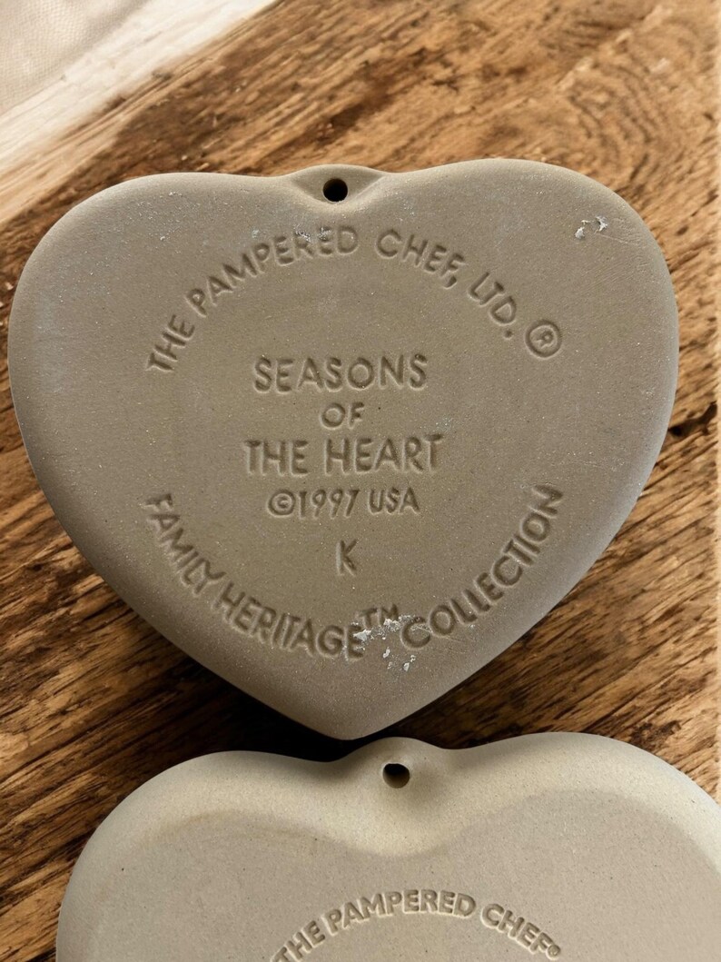 Puede incluir: Dos adornos de cer&aacute;mica en forma de coraz&oacute;n, de color marr&oacute;n claro. El texto en el adorno superior dice "THE PAMPERED CHEF, LTD. SEASONS OF THE HEART &copy;1997 USA FAMILY HERITAGE COLLECTION". Cada adorno tiene un peque&ntilde;o agujero en la parte superior para colgar.