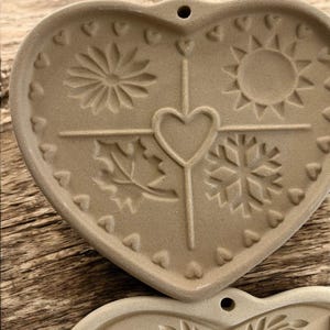 Puede incluir: Adorno de cer&aacute;mica en forma de coraz&oacute;n de color beige con dise&ntilde;os de temporada en relieve. El adorno presenta una flor, un sol, una hoja y un copo de nieve, con un coraz&oacute;n en el centro. Peque&ntilde;os corazones bordean el borde. Un agujero est&aacute; en la parte superior para colgar.