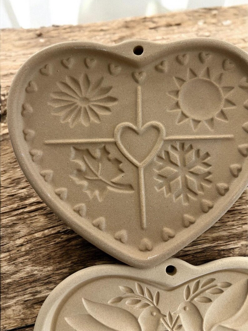 Puede incluir: Adorno de cer&aacute;mica en forma de coraz&oacute;n de color beige con dise&ntilde;os de temporada en relieve. El adorno presenta una flor, un sol, una hoja y un copo de nieve, con un coraz&oacute;n en el centro. Peque&ntilde;os corazones bordean el borde. Un agujero est&aacute; en la parte superior para colgar.