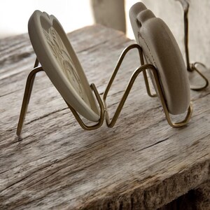 Puede incluir: Dos platos decorativos de cer&aacute;mica con dise&ntilde;os en relieve se exhiben en soportes de metal color lat&oacute;n. Los platos son de color blanquecino y tienen forma de coraz&oacute;n. Los soportes est&aacute;n sobre una superficie de madera envejecida.