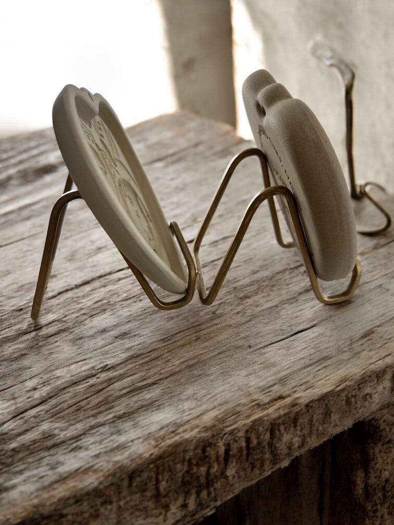 Puede incluir: Dos platos decorativos de cer&aacute;mica con dise&ntilde;os en relieve se exhiben en soportes de metal color lat&oacute;n. Los platos son de color blanquecino y tienen forma de coraz&oacute;n. Los soportes est&aacute;n sobre una superficie de madera envejecida.