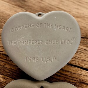 Puede incluir: Dos utensilios de cocina grises en forma de coraz&oacute;n con un agujero en la parte superior para colgar. El texto "GARDENS OF THE HEART" y "THE PAMPERED CHEF LTD." est&aacute;n grabados en la parte delantera, junto con "1996 U.S.A."