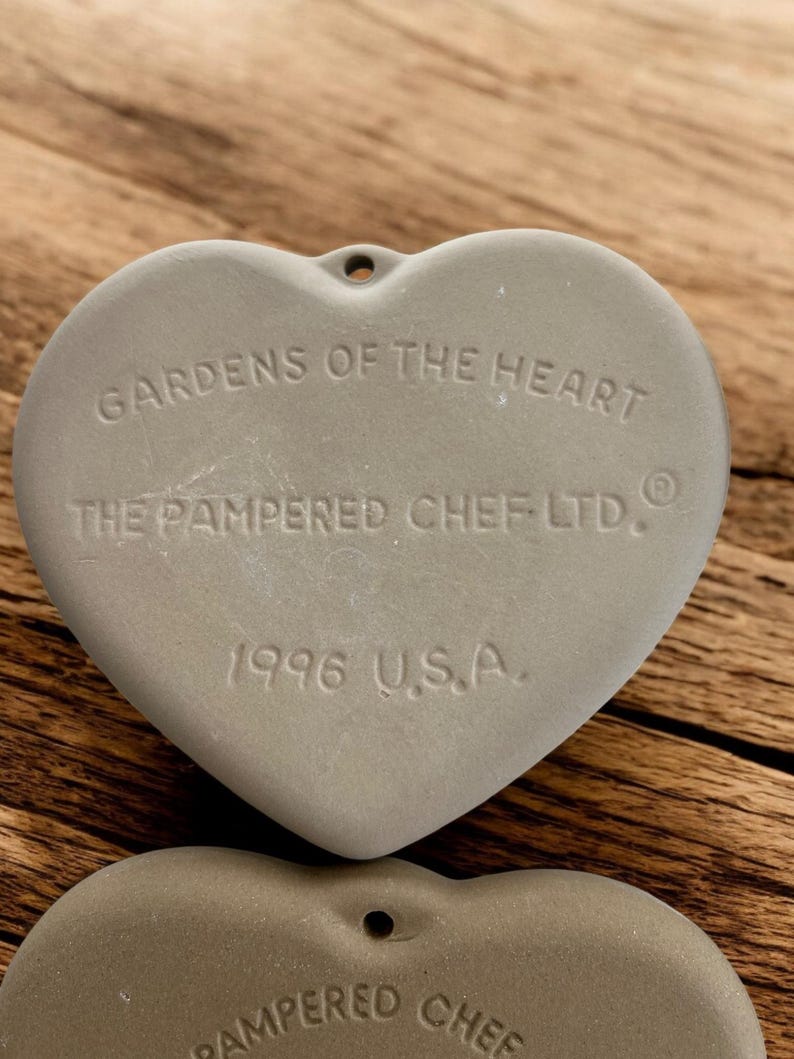 Puede incluir: Dos utensilios de cocina grises en forma de coraz&oacute;n con un agujero en la parte superior para colgar. El texto "GARDENS OF THE HEART" y "THE PAMPERED CHEF LTD." est&aacute;n grabados en la parte delantera, junto con "1996 U.S.A."
