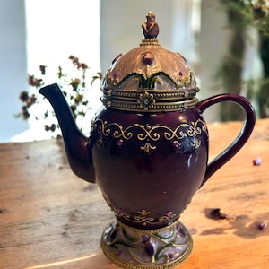 May include: A decorative, ornate teapot in a deep burgundy color with gold accents. The lid is tan with purple and green floral designs. The teapot has a curved handle and spout, and sits on a decorative base. A small floral finial tops the lid.