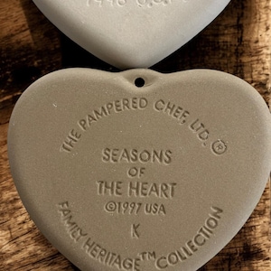 Puede incluir: Dos adornos de cer&aacute;mica en forma de coraz&oacute;n, de color claro. El adorno superior est&aacute; estampado con "1996 U.S.A.". El adorno inferior dice "The Pampered Chef, Ltd." y "Seasons of the Heart &copy;1997 USA Family Heritage Collection."