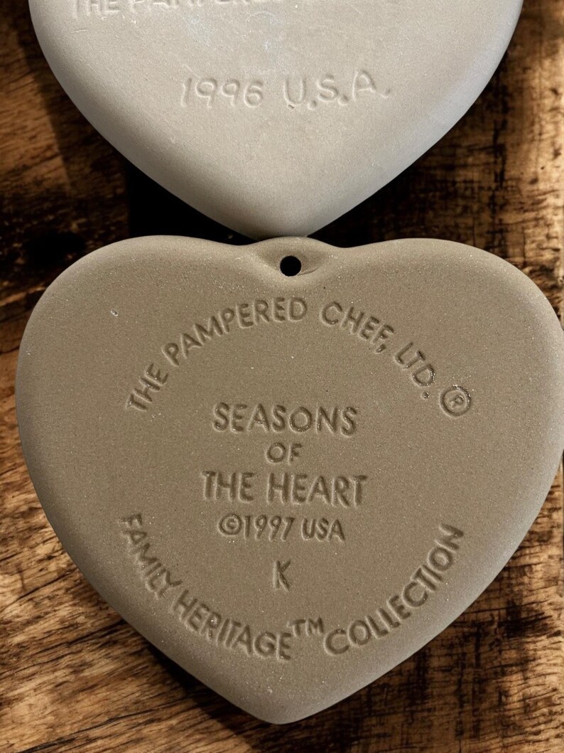 Puede incluir: Dos adornos de cer&aacute;mica en forma de coraz&oacute;n, de color claro. El adorno superior est&aacute; estampado con "1996 U.S.A.". El adorno inferior dice "The Pampered Chef, Ltd." y "Seasons of the Heart &copy;1997 USA Family Heritage Collection."