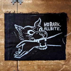 May include: Black fabric patch featuring a white illustration of a wolf with the text "NO BARK, ALL BITE." The wolf is drawn in a cartoon style, with visible teeth and a threatening look. The patch has a raw edge and is likely handmade.