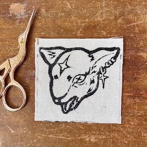 May include: A square, off-white fabric patch with a black linocut print of a stylized cat's head. The cat has star-shaped accents and a detailed ear. Gold scissors are visible in the background.