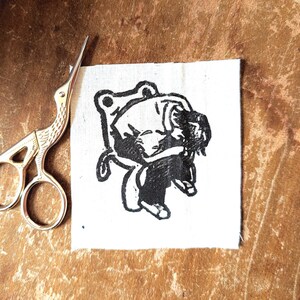 May include: A black and white linocut print of a figure on a square of white fabric. The figure is stylized and appears to be sitting or crouching. A pair of gold-coloured scissors is positioned to the left of the fabric square.