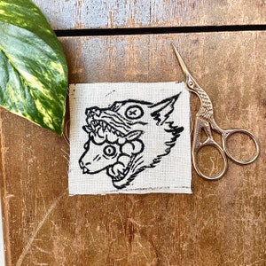 May include: Black and white embroidered patch of a wolf's head with a floral design on a white fabric background.