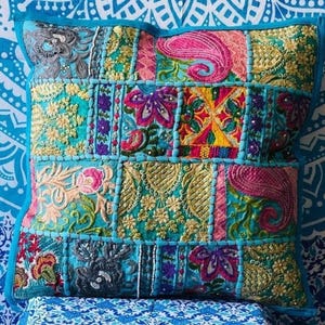 May include: Square cushion with a patchwork design. The cushion features a variety of colourful embroidered patterns in shades of teal, pink, gold, and purple. The cushion has a teal border.