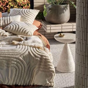 May include: A cream-coloured bedding set featuring a raised, tufted arch pattern on the duvet cover and matching throw pillows. The set is paired with a small, speckled, cone-shaped side table and a large, smooth, dark grey rounded planter.