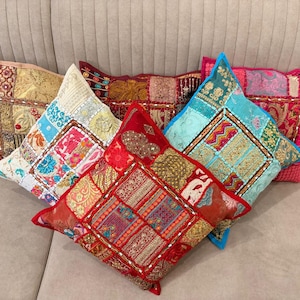 Puede incluir: Cinco cojines cuadrados con un diseño de patchwork de varias telas, bordados intrincados, detalles en hilo metálico y pequeños adornos de espejo. Los cojines muestran una paleta de colores vibrantes que incluye rojo, dorado, azul y rosa.