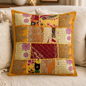 May include: A square throw pillow featuring a patchwork design of various fabrics in shades of yellow, orange, red, and gold. The surface displays embroidered floral patterns, paisley motifs, and decorative stitching in a bright yellow thread.