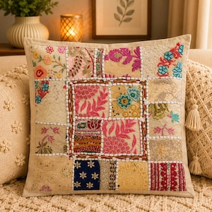 Op de afbeelding: Een vierkant sierkussen met een patchworkontwerp van diverse geborduurde stoffen panelen in tinten roze, blauw, geel en crème. Het oppervlak is versierd met ingewikkelde bloemmotieven, lovertjes en kralenwerk, waarbij dikke witte stiksels elk afzonderlijk rechthoekig en vierkant gedeelte omlijnen.