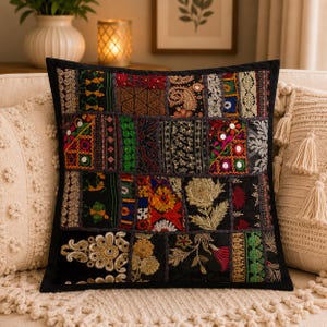 Può includere: Un cuscino quadrato caratterizzato da un design patchwork composto da vari pannelli in tessuto nero. I pannelli sono decorati con ricami complessi, filo dorato metallizzato, motivi floreali colorati, piccoli specchi circolari e forme geometriche.
