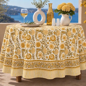 Può includere: Una tovaglia rotonda caratterizzata da un motivo floreale stampato a blocchi color giallo senape e verde oliva su fondo bianco, rifinita con un bordo a righe. Il tessuto copre un tavolo circolare, creando una copertura tessile decorativa.