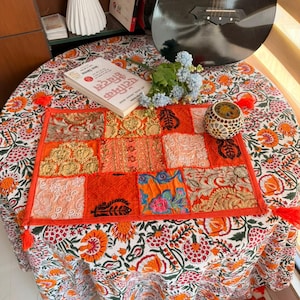 Könnte beinhalten: Ein runder Tisch mit einer floral gemusterten Tischdecke. Ein Patchwork-Tischset in Orange- und Goldtönen liegt auf dem Tisch. Ein Buch, eine Vase und dekorative Gegenstände sind ebenfalls vorhanden. Eine Gitarre lehnt an der Wand.
