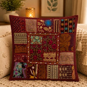 Peut inclure: Un coussin carré composé d'un assemblage de panneaux de tissu rouge foncé, bordeaux et marron. La surface est ornée de broderies complexes dorées, bleues et vertes, de motifs floraux et de détails en fils métalliques.