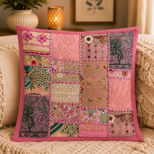 May include: A square throw cushion featuring a patchwork design of various pink, purple, and green fabric panels. The surface is decorated with intricate embroidery, floral patterns, and metallic thread accents, all framed by a solid pink border.